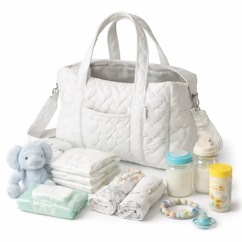 Sac à langer blanc matelassé vide posé sur fond blanc, entouré de couches, lingettes, biberons, lange, tétine et doudou pour illustrer la capacité de rangement.