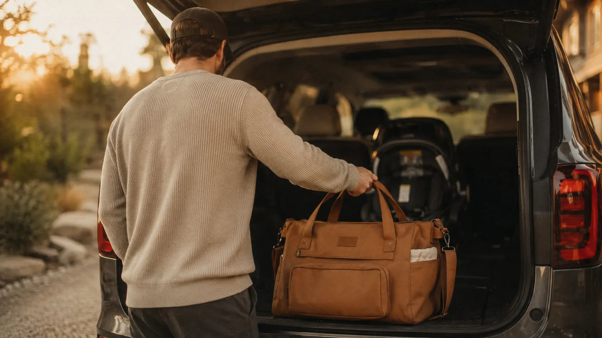 Père de dos chargeant un grand sac à langer caramel dans le coffre d'une voiture, siège bébé visible en arrière-plan, lumière dorée de fin de journée.