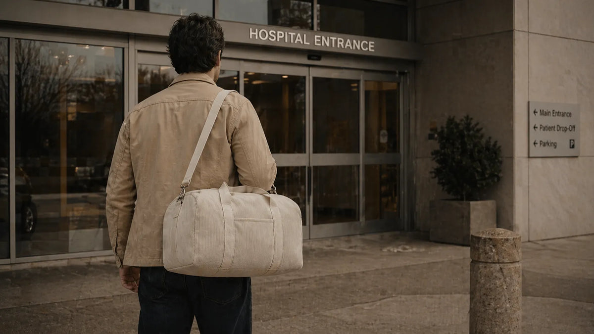 Père de dos devant l'entrée d'une maternité, tenant un grand sac à langer beige en bandoulière.