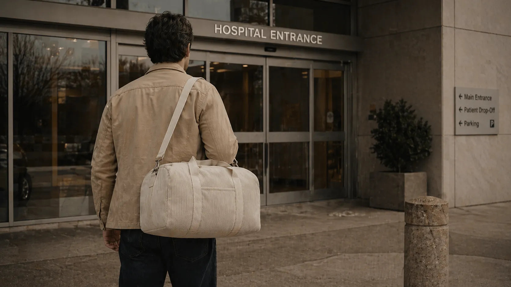 Père de dos devant l'entrée d'une maternité, tenant un grand sac à langer beige en bandoulière.