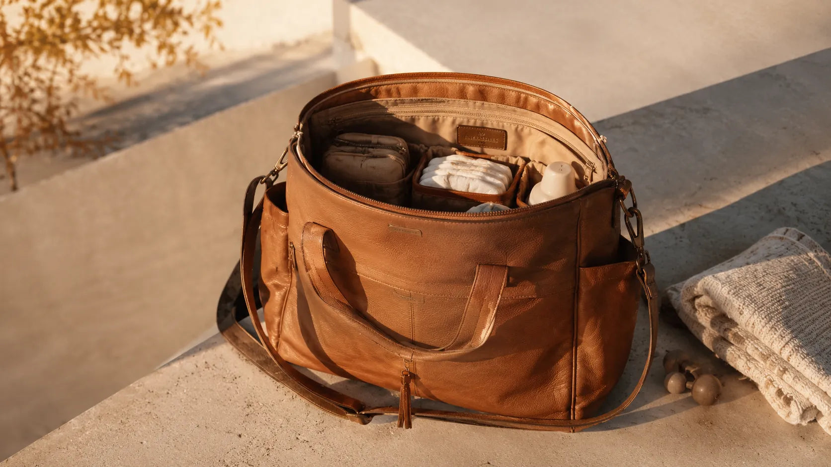 Sac à langer effet cuir cognac légèrement ouvert posé sur une surface en béton clair, compartiments intérieurs visibles, lumière dorée latérale, grain pellicule. Image