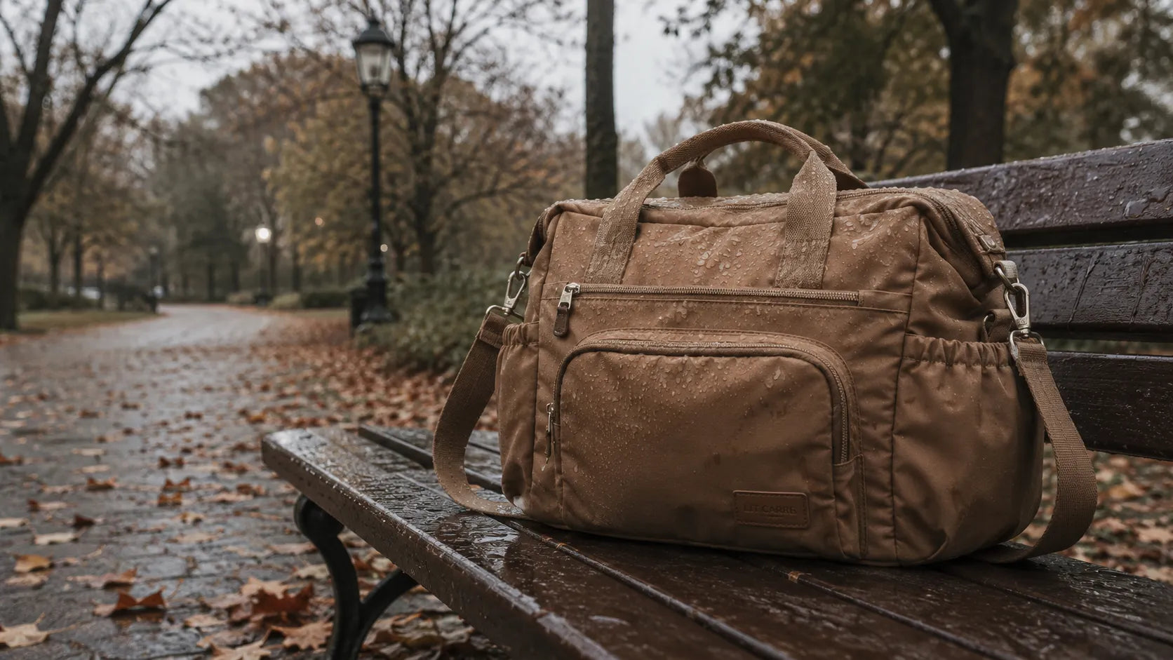 Sac à langer caramel imperméable posé sur un banc mouillé dans un parc sous la pluie, gouttes d'eau visibles sur la surface du sac.