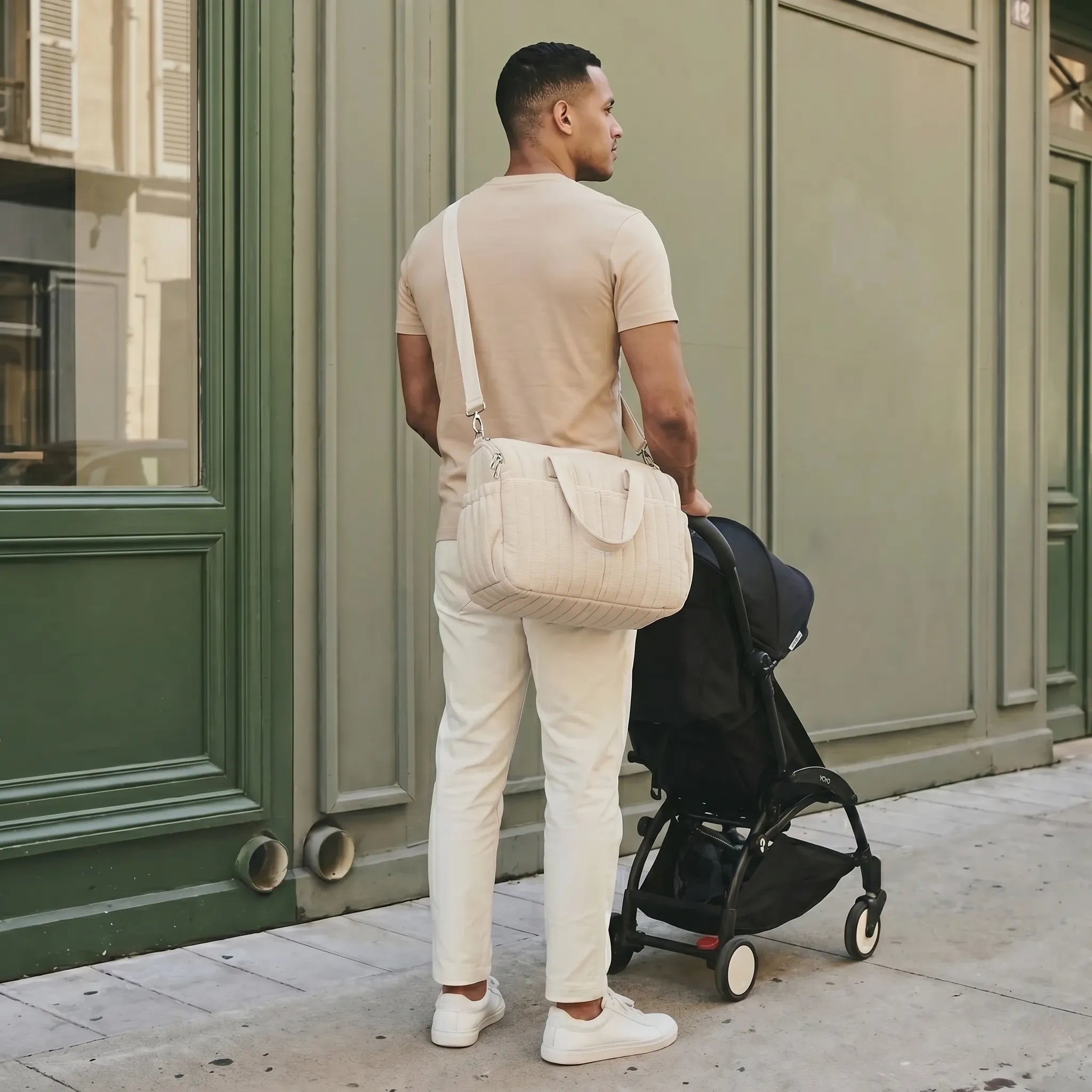 Homme de dos portant un sac à langer crème matelassé en bandoulière dans une rue  parisienne avec façade verte haussmannienne, poussant une poussette Babyzen YOYO —  scène lifestyle père moderne