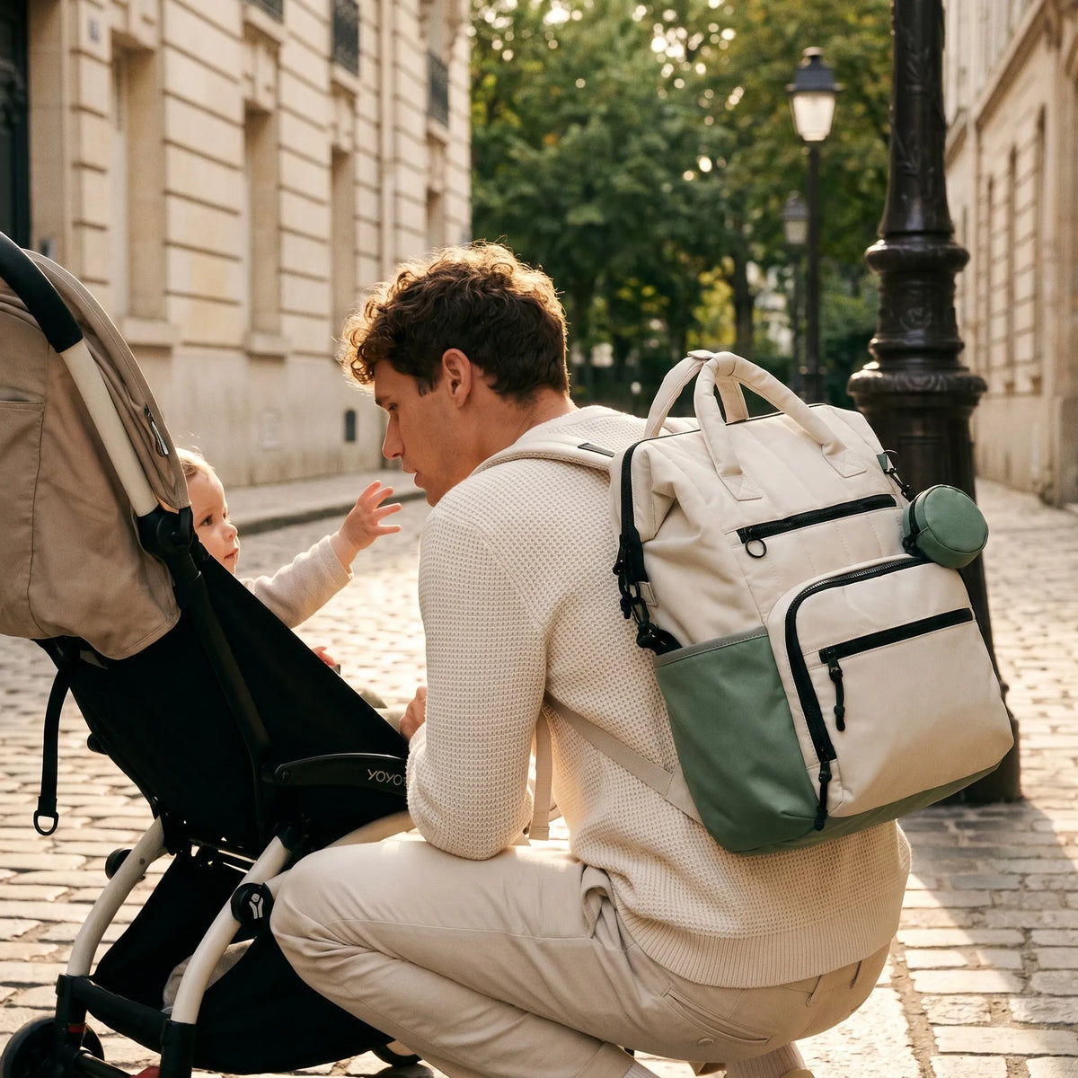 Homme de dos en mouvement portant un sac à dos à langer matelassé crème 
et vert sauge, poussant une poussette Babyzen YOYO noire dans une rue 
parisienne animée au petit matin — scène lifestyle dynamique Image