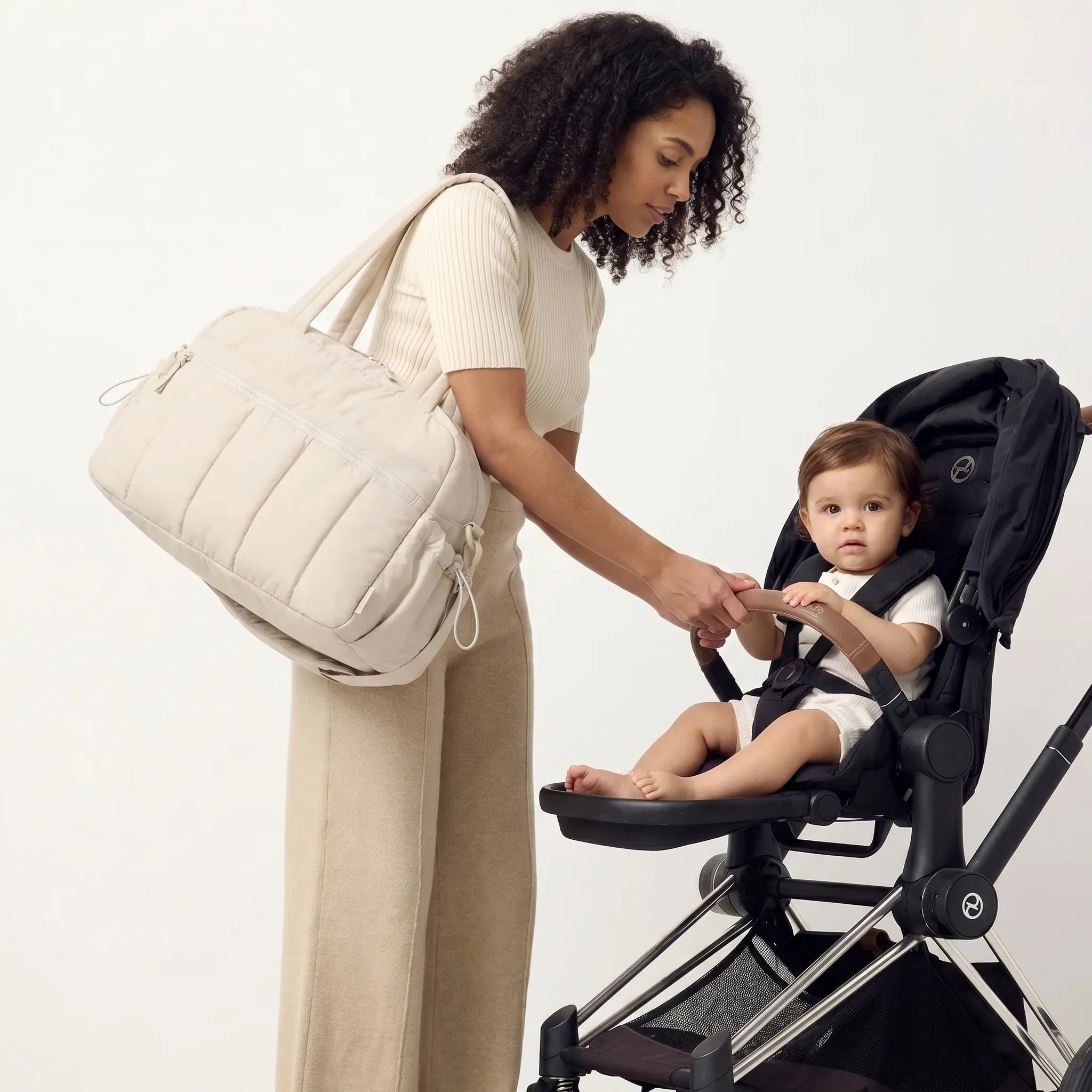 Femme portant un sac à langer duffle beige matelassé en bandoulière se penchant vers son bébé dans une poussette Cybex