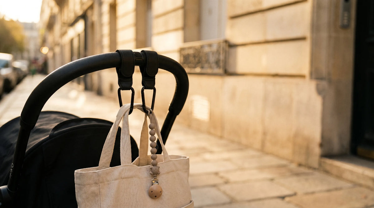 Crochets attache poussette noirs effet cuir fixés sur le guidon d'une poussette 
Babyzen YOYO, sac suspendu et attache-tétine en silicone taupe, dans une rue 
parisienne au coucher du soleil — bannière collection accessoires