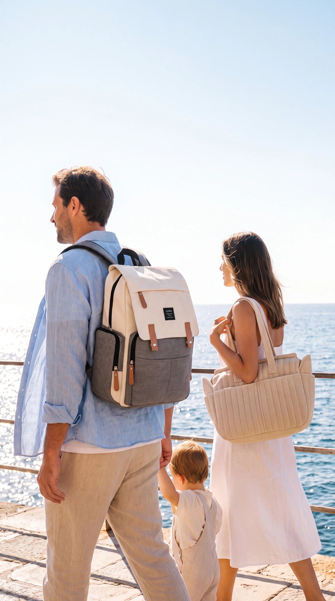 Famille de dos marchant sur une promenade méditerranéenne ensoleillée, père 
portant un sac à dos canvas bicolore crème et gris LAND bien visible, mère 
avec tote beige côtelé, enfant entre eux — bannière mobile lifestyle été