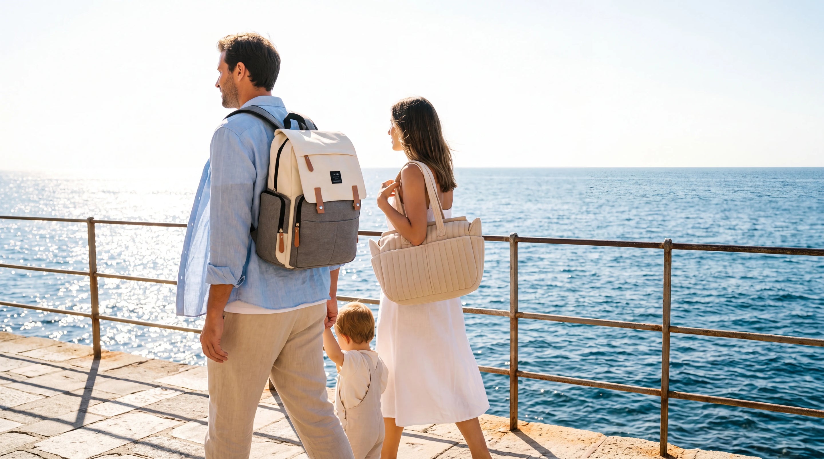 Couple de jeunes parents en tenue estivale portant un sac à dos canvas bicolore 
crème et gris et un sac tote beige côtelé en coton, promenade en bord de mer 
méditerranéen en été — bannière lifestyle collection sacs à langer