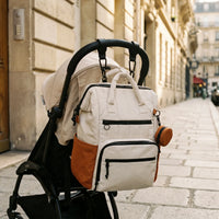 Deux crochets attaches poussette noirs effet cuir fixés sur guidon de poussette 
Babyzen YOYO noire, sac à dos à langer crème et orange bicolore suspendu — 
scène lifestyle rue parisienne dorée