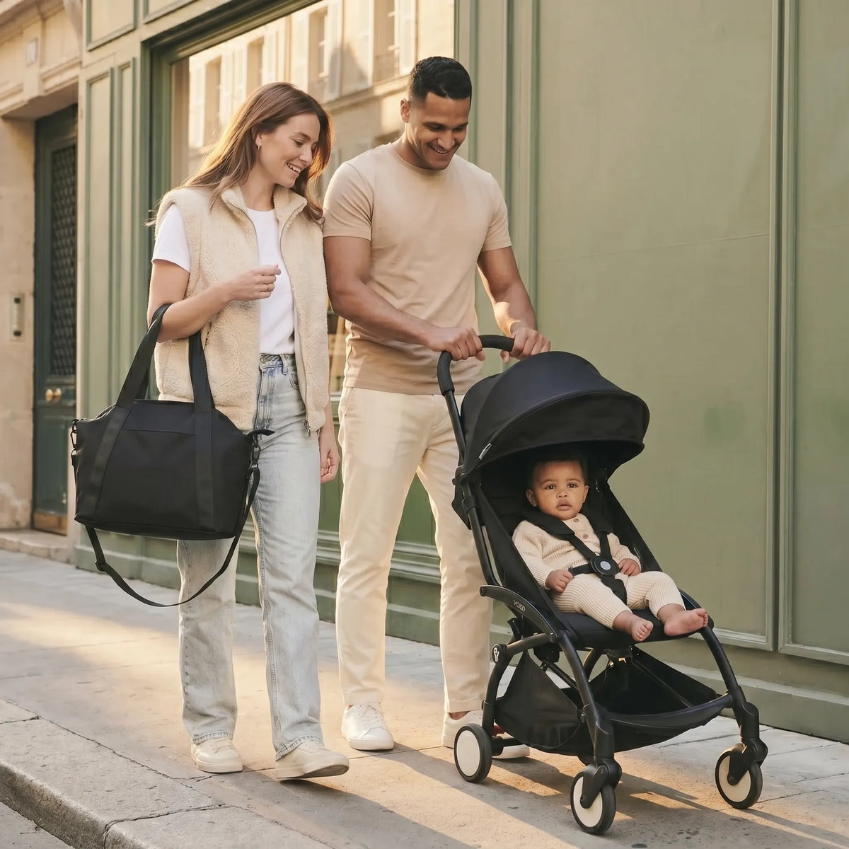 Jeune couple souriant marchant dans une rue parisienne, la maman portant un sac à langer bandoulière noir et le papa poussant une poussette compacte Yoyo avec leur bébé