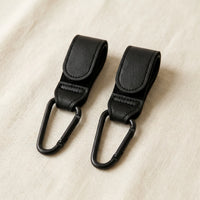 Deux crochets attaches poussette noirs effet cuir avec sangle velcro réglable 
et mousqueton à clip matte black, posés sur surface lin crème — 
photo lifestyle produit fond neutre