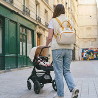 Jeune maman de dos portant un sac à dos à langer bicolore écru avec finitions moutarde tout en poussant une poussette avec son bébé dans une rue parisienne