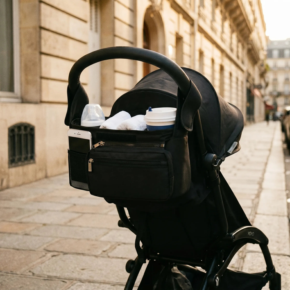 Organisateur de poussette noir universel avec porte-biberons, poche téléphone 
mesh et poche avant zippée, monté sur guidon d'une poussette Babyzen YOYO 
noire — scène lifestyle rue parisienne dorée