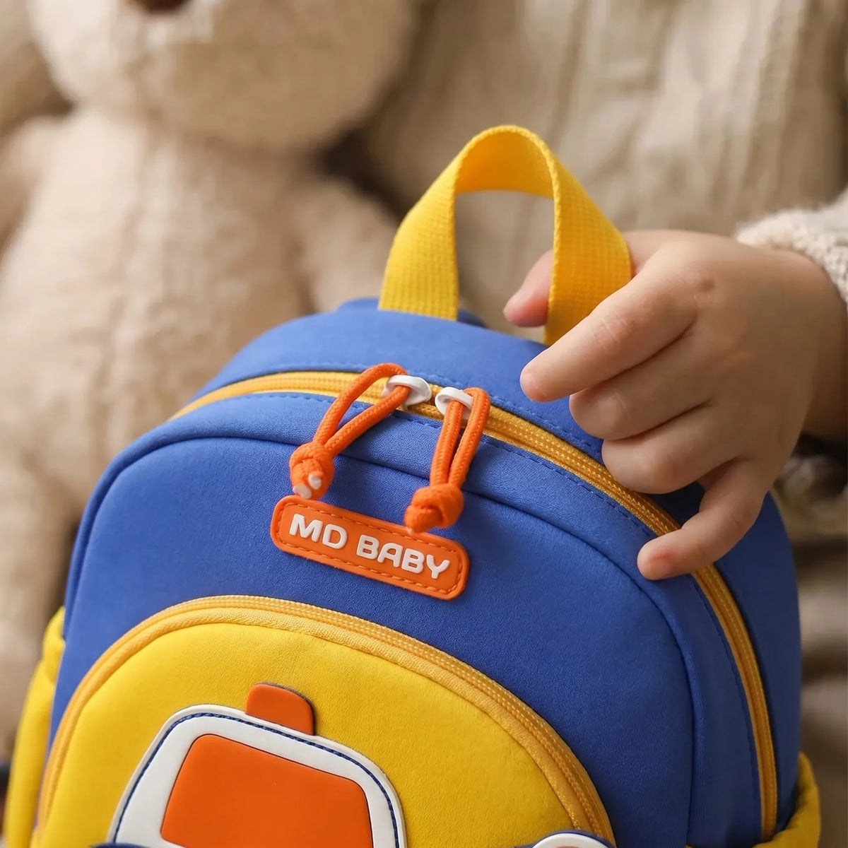 Gros plan sur la main d'un jeune enfant qui ouvre le zip à tirette orange du sac à dos crèche 1-3 ans bleu et jaune avec doudou en arrière-plan