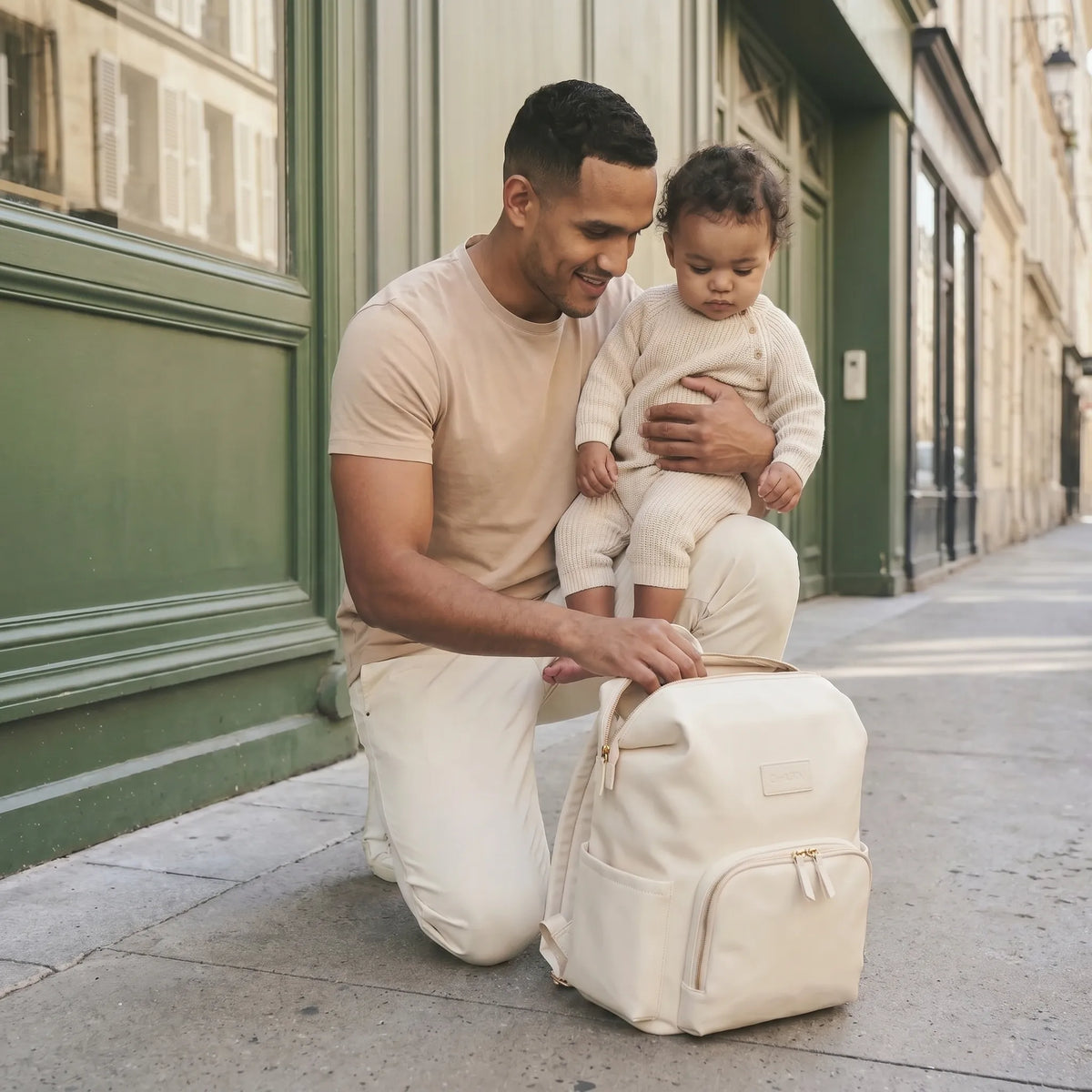 Jeune papa accroupi tenant son bébé dans les bras et ouvrant un sac à dos à langer effet cuir écru posé sur le trottoir d'une rue parisienne