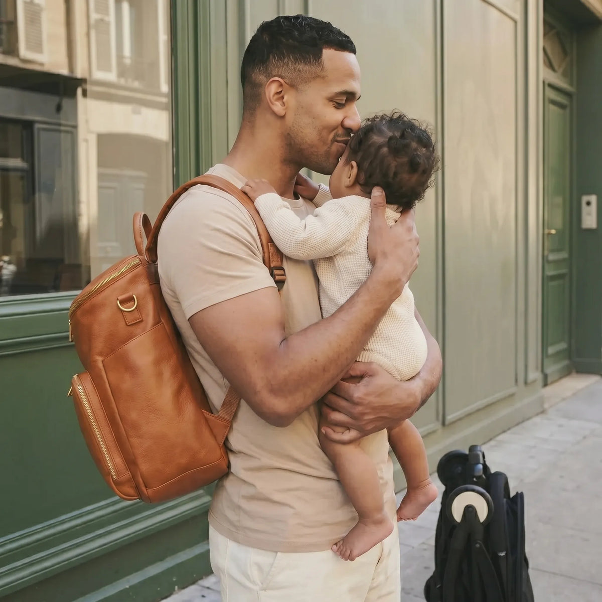 Papa portant un sac à dos à langer en cuir PU camel sur le dos et tenant son bébé dans les bras en l'embrassant tendrement sur le front dans une rue parisienne avec poussette à côté