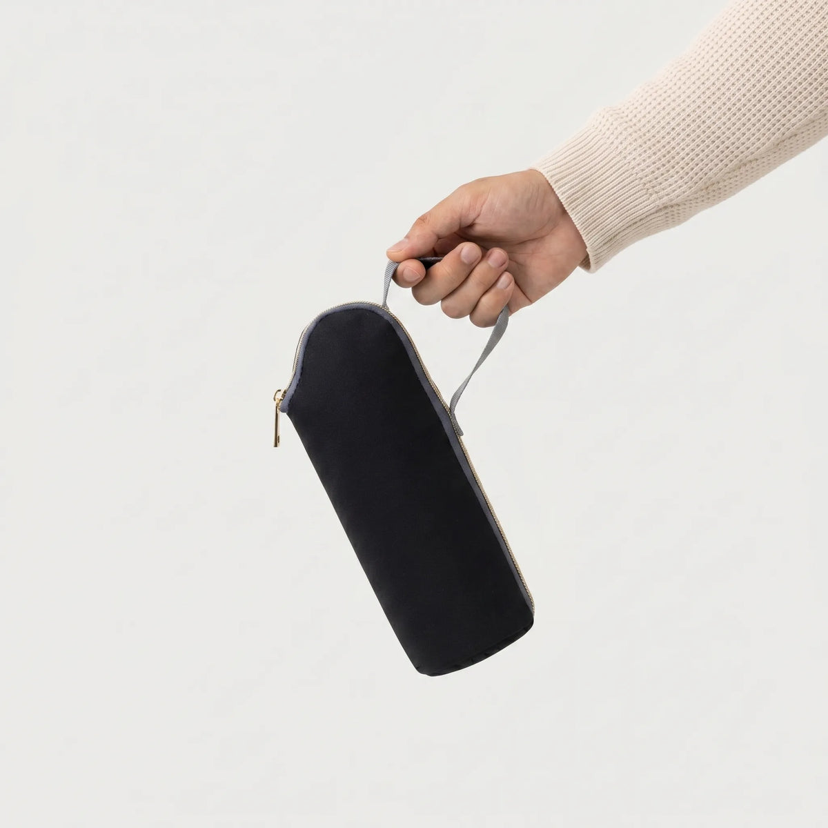 Main tenant une pochette isotherme biberon noire avec zip doré et anse grise, 
manche pull crème waffle knit visible — photo lifestyle produit studio fond blanc
