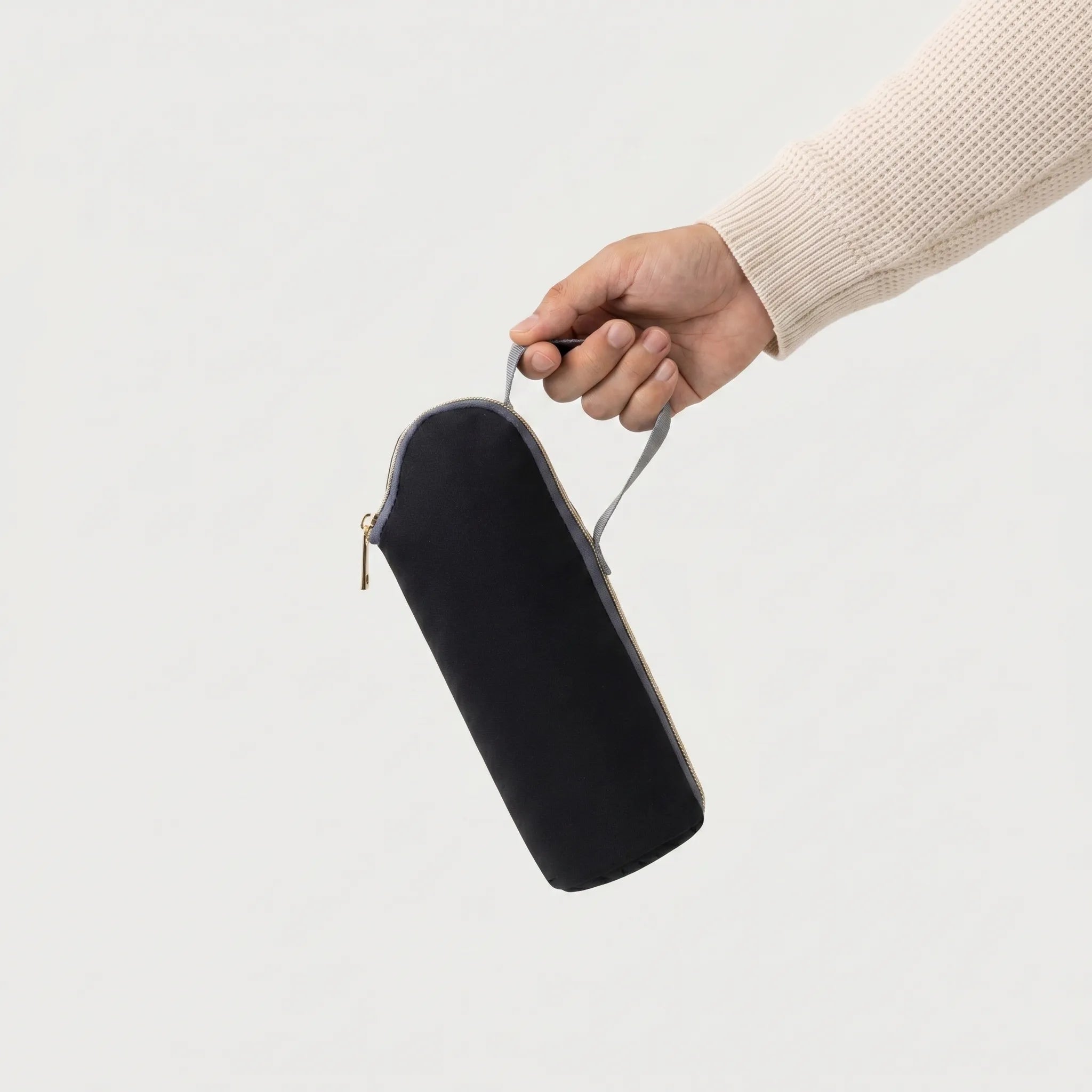 Main tenant une pochette isotherme biberon noire avec zip doré et anse grise, 
manche pull crème waffle knit visible — photo lifestyle produit studio fond blanc