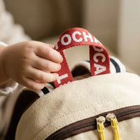 Gros plan d'une main d'enfant saisissant la poignée de transport rouge brodée Tochaso d'un sac à dos en canvas beige avec zips jaunes