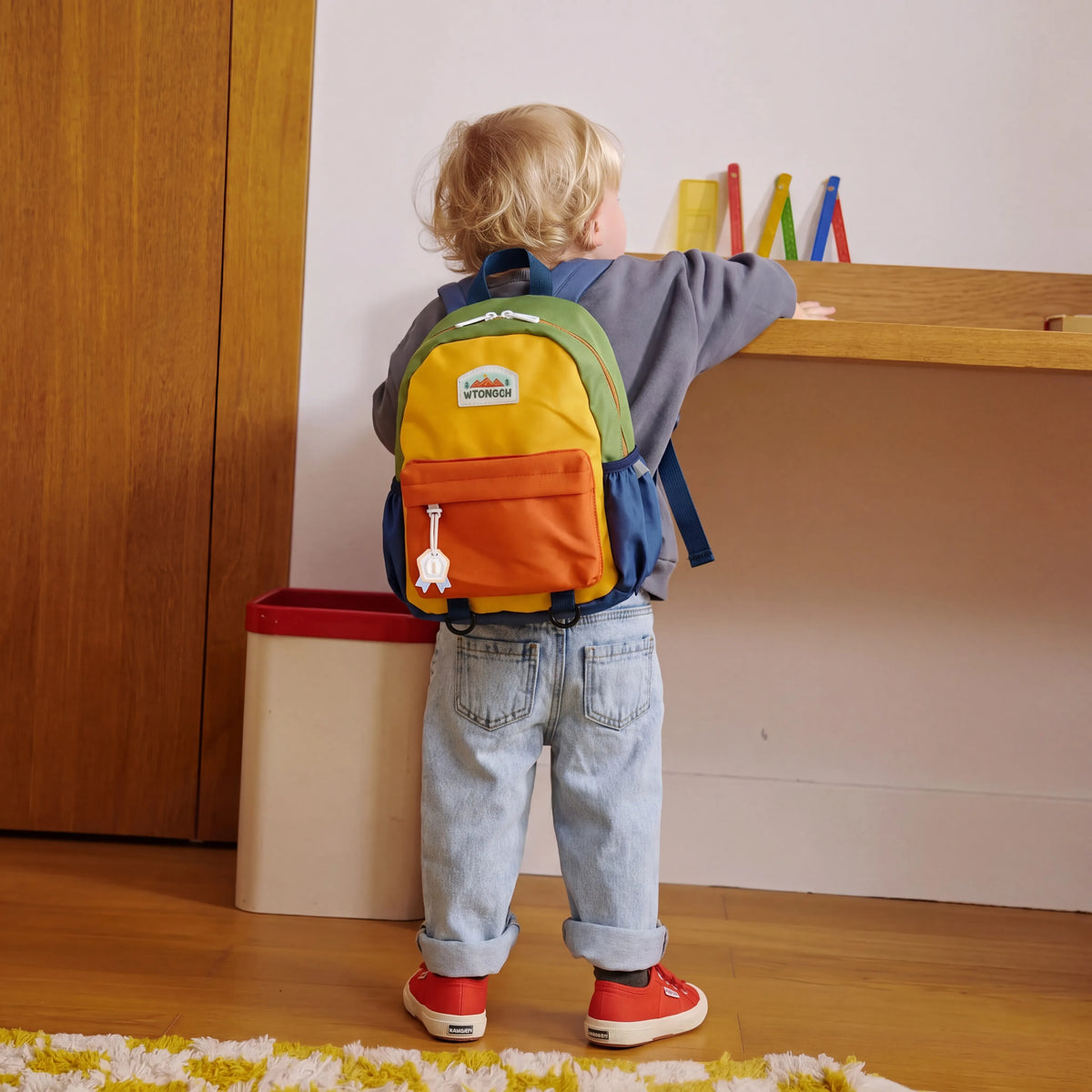 Enfant de dos portant un sac à dos crèche multicolore vert jaune et orange dans une chambre
