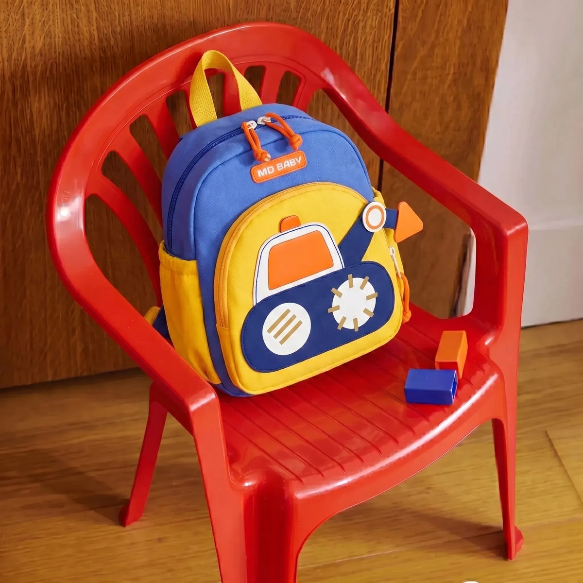 Sac à dos crèche 1-3 ans bleu et jaune avec motif engin de chantier en relief posé sur une chaise enfant rouge avec cubes de motricité