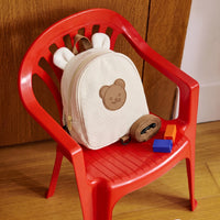 Sac à dos crèche beige en toile canvas avec oreilles et écusson ourson posé sur une chaise enfant rouge avec sangle de sécurité et cubes de motricité