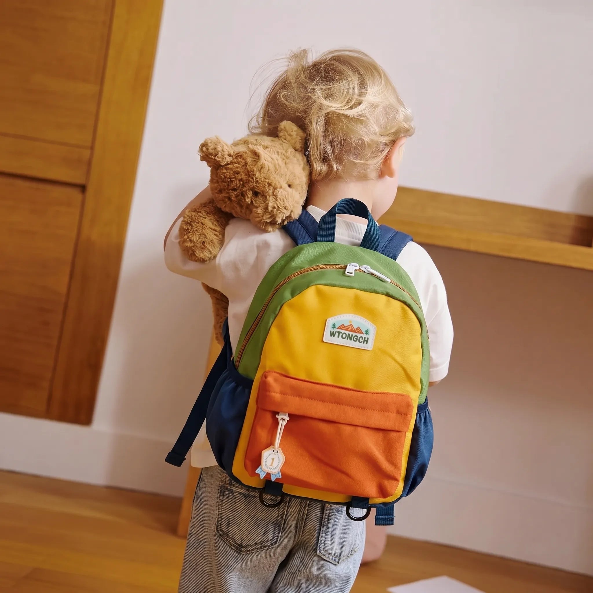 Enfant de dos portant un sac à dos crèche multicolore vert jaune et orange tenant un ours en peluche dans une chambre