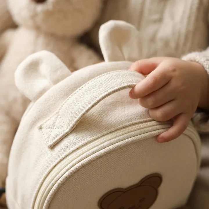 Gros plan d'une main d'enfant tenant la poignée d'un petit sac à dos rond en canvas beige en forme d'ours avec oreilles, posé devant une peluche teddy bear