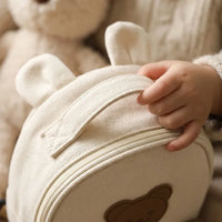Gros plan d'une main d'enfant tenant la poignée d'un petit sac à dos rond en canvas beige en forme d'ours avec oreilles, posé devant une peluche teddy bear
