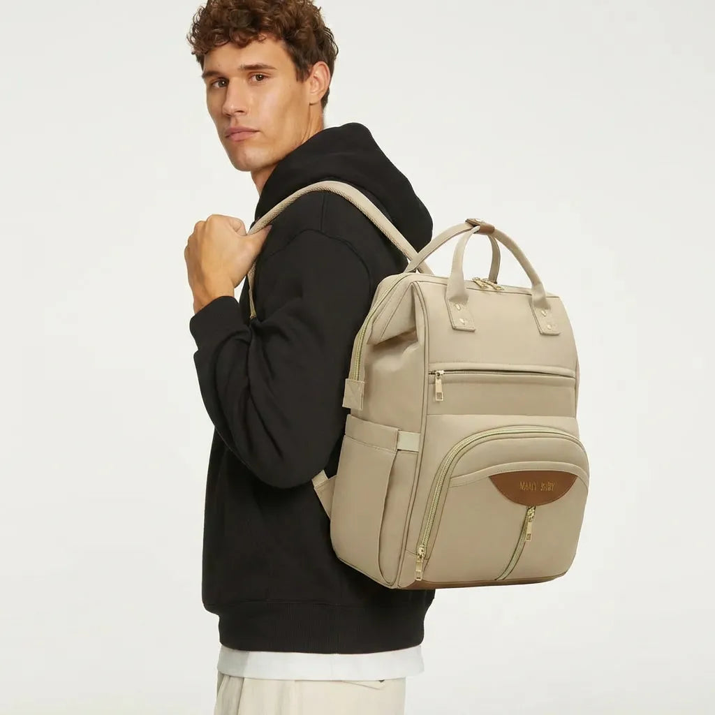 Homme portant un sac à dos à langer beige avec détails dorés et patch marron sur fond blanc