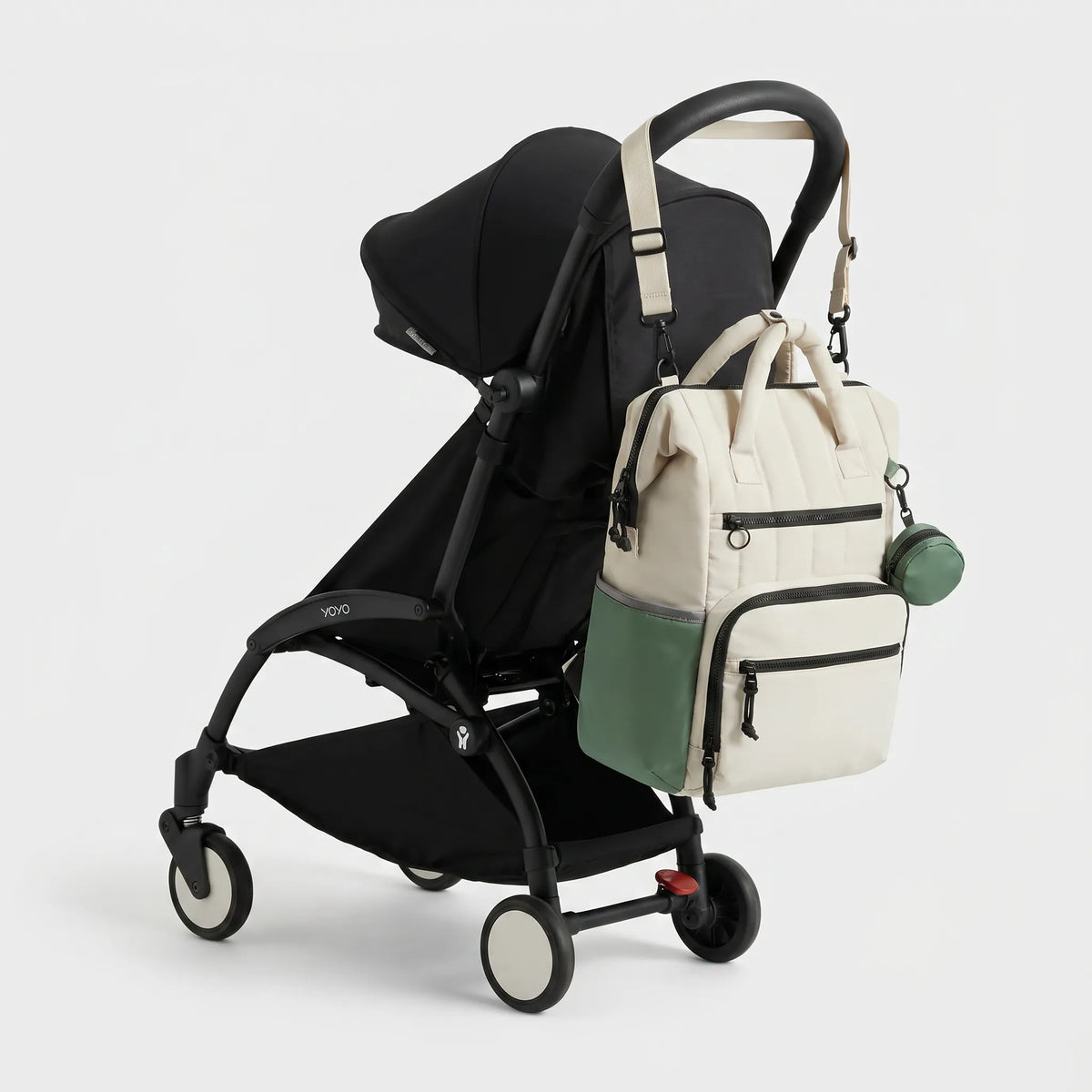 Sac à dos à langer bicolore crème et vert sauge avec matelassage vertical et 
mini pochette porte-sucette, suspendu au guidon d'une poussette Babyzen YOYO 
noire — vue lifestyle studio