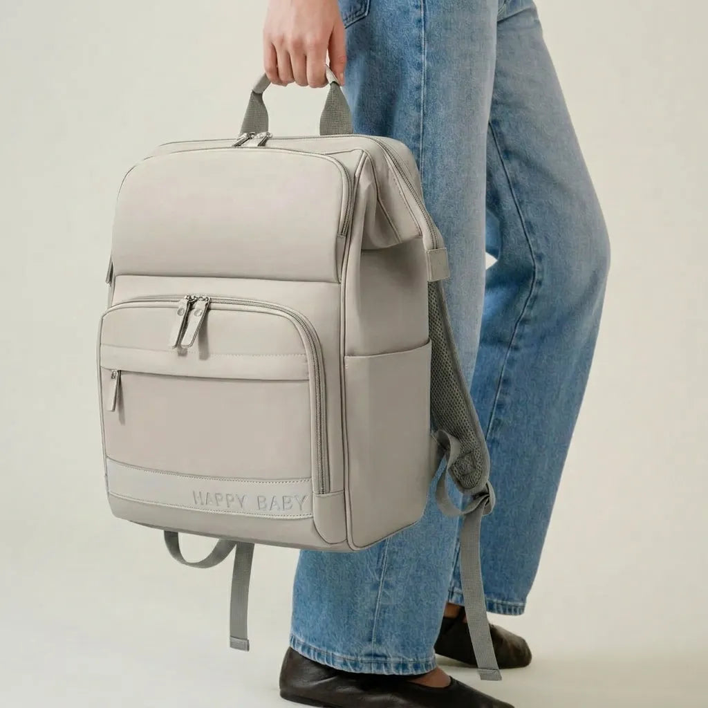 Personne tenant un sac à dos à langer beige structuré par la poignée supérieure, bretelles tombant naturellement sur les côtés