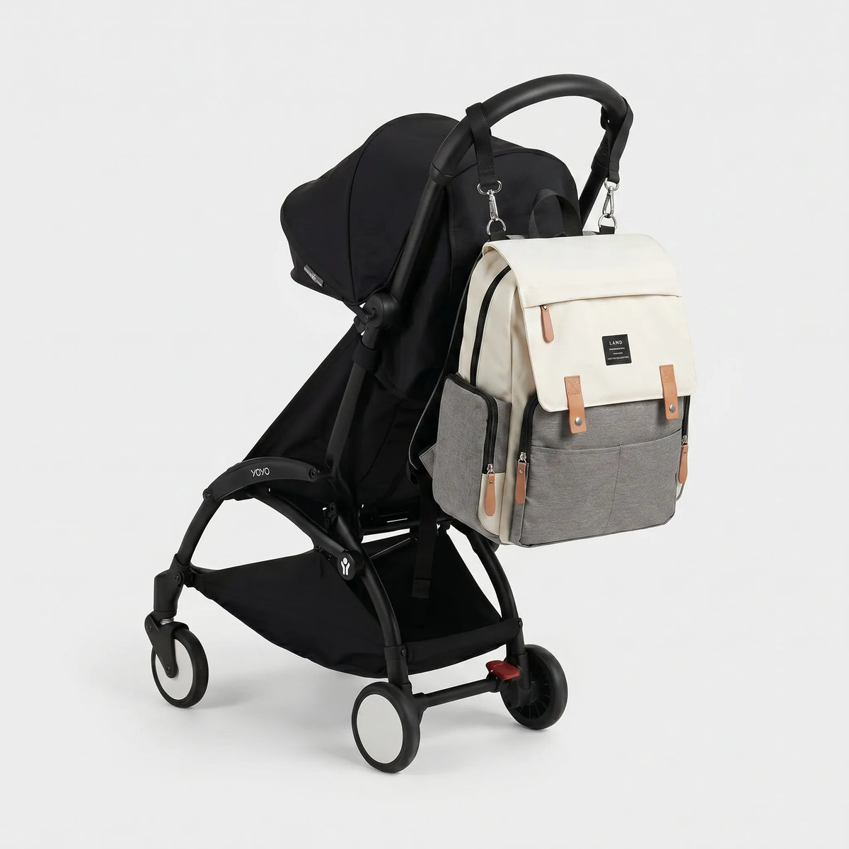 Sac à dos à langer bicolore crème et gris chiné avec rabat canvas, sangles cuir 
caramel et badge LAND, suspendu au guidon d'une poussette Babyzen YOYO noire — 
vue lifestyle studio