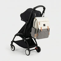 Sac à dos à langer bicolore crème et gris chiné avec rabat canvas, sangles cuir 
caramel et badge LAND, suspendu au guidon d'une poussette Babyzen YOYO noire — 
vue lifestyle studio