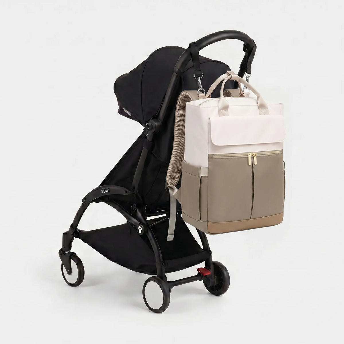 Sac à dos à langer bicolore crème et taupe avec hardware doré et poignées larges, 
suspendu au guidon d'une poussette Babyzen YOYO noire — vue lifestyle studio