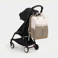 Sac à dos à langer bicolore crème et taupe avec hardware doré et poignées larges, 
suspendu au guidon d'une poussette Babyzen YOYO noire — vue lifestyle studio