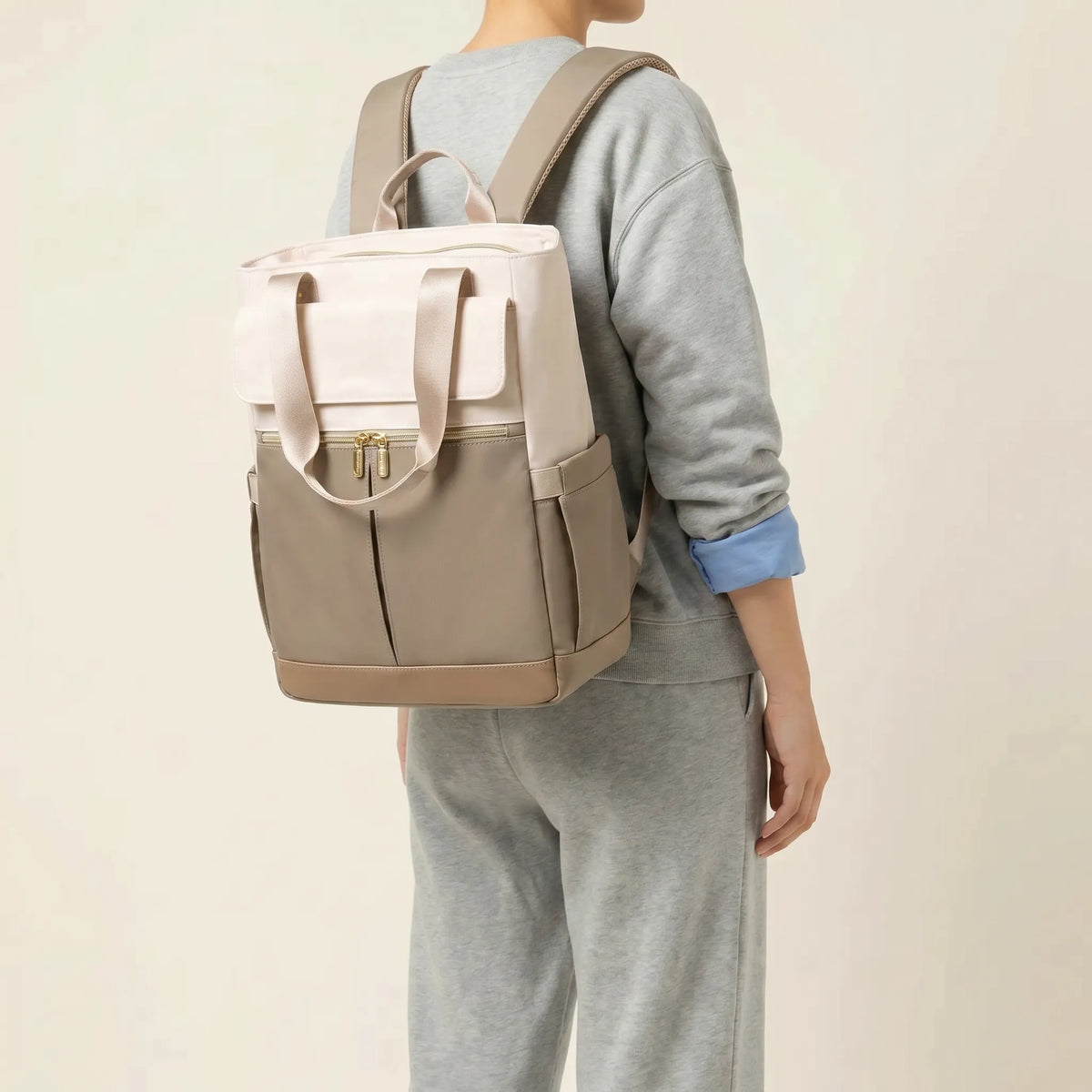 Personne de dos portant un sac à dos à langer bicolore crème et taupe avec bretelles 
ergonomiques maillées et hardware doré — vue lifestyle studio fond blanc chaud
