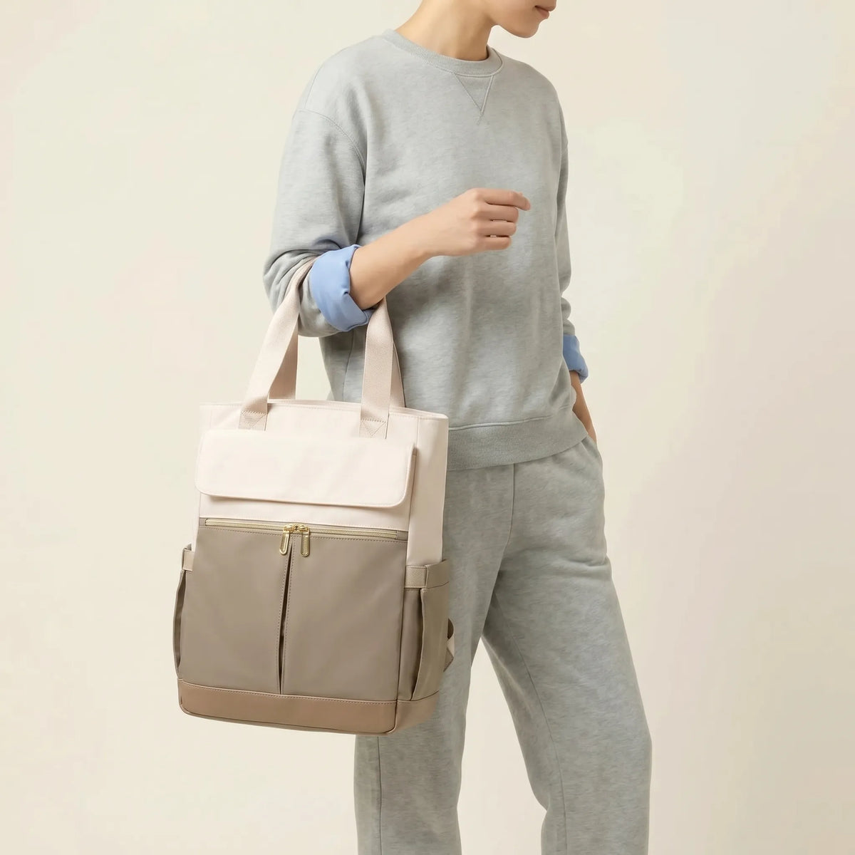 Personne tenant à la main un sac à dos à langer bicolore crème et taupe avec 
hardware doré et poignées larges — vue lifestyle studio fond blanc chaud