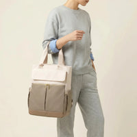 Personne tenant à la main un sac à dos à langer bicolore crème et taupe avec 
hardware doré et poignées larges — vue lifestyle studio fond blanc chaud