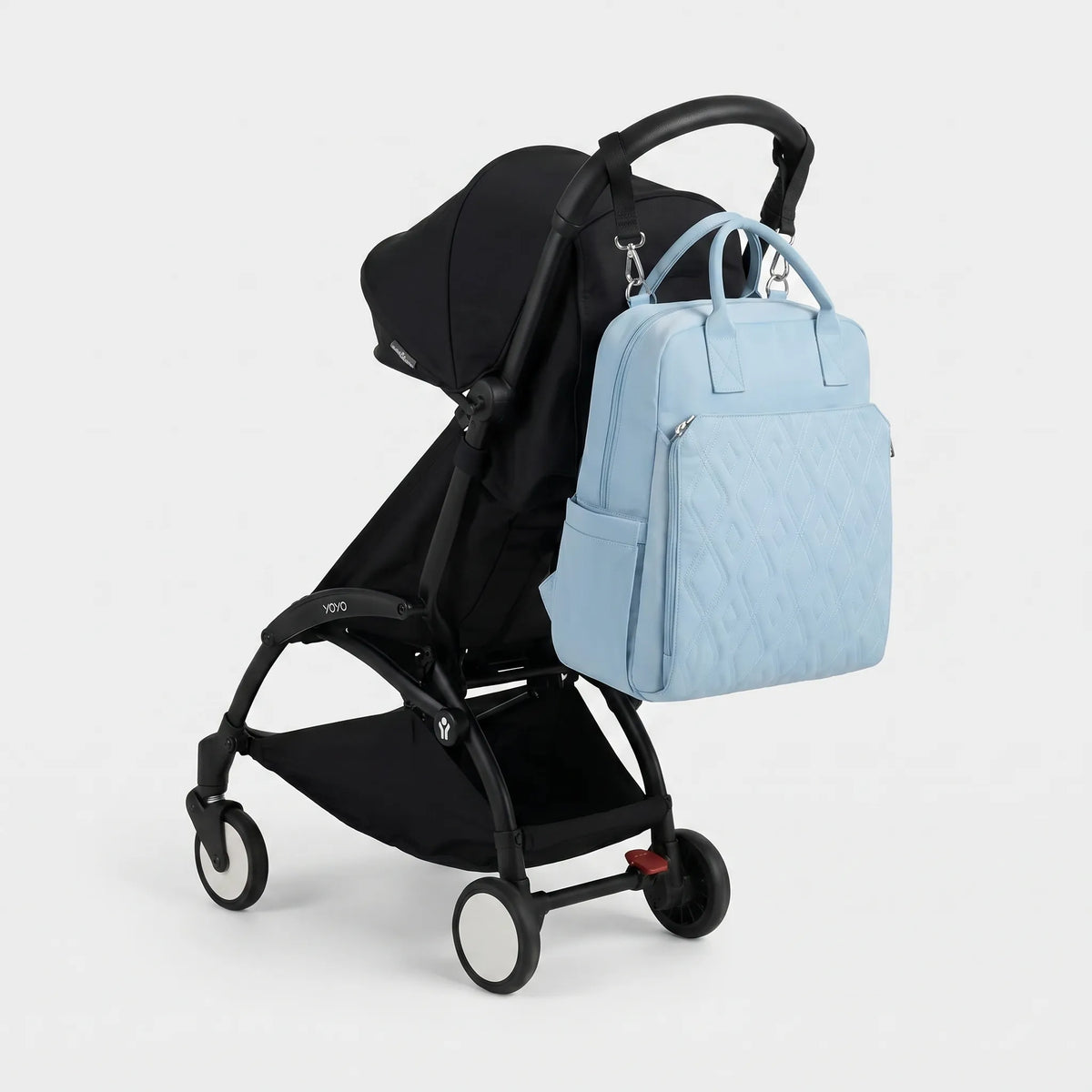 Sac à dos à langer bleu ciel avec broderie diamant en relief et hardware argenté, 
suspendu au guidon d'une poussette Babyzen YOYO noire — vue lifestyle studio