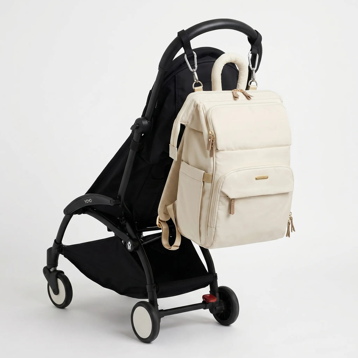 Sac à dos à langer monochrome crème et hardware doré avec poignée tubulaire 
rembourrée, suspendu au guidon d'une poussette Babyzen YOYO noire — 
vue lifestyle studio