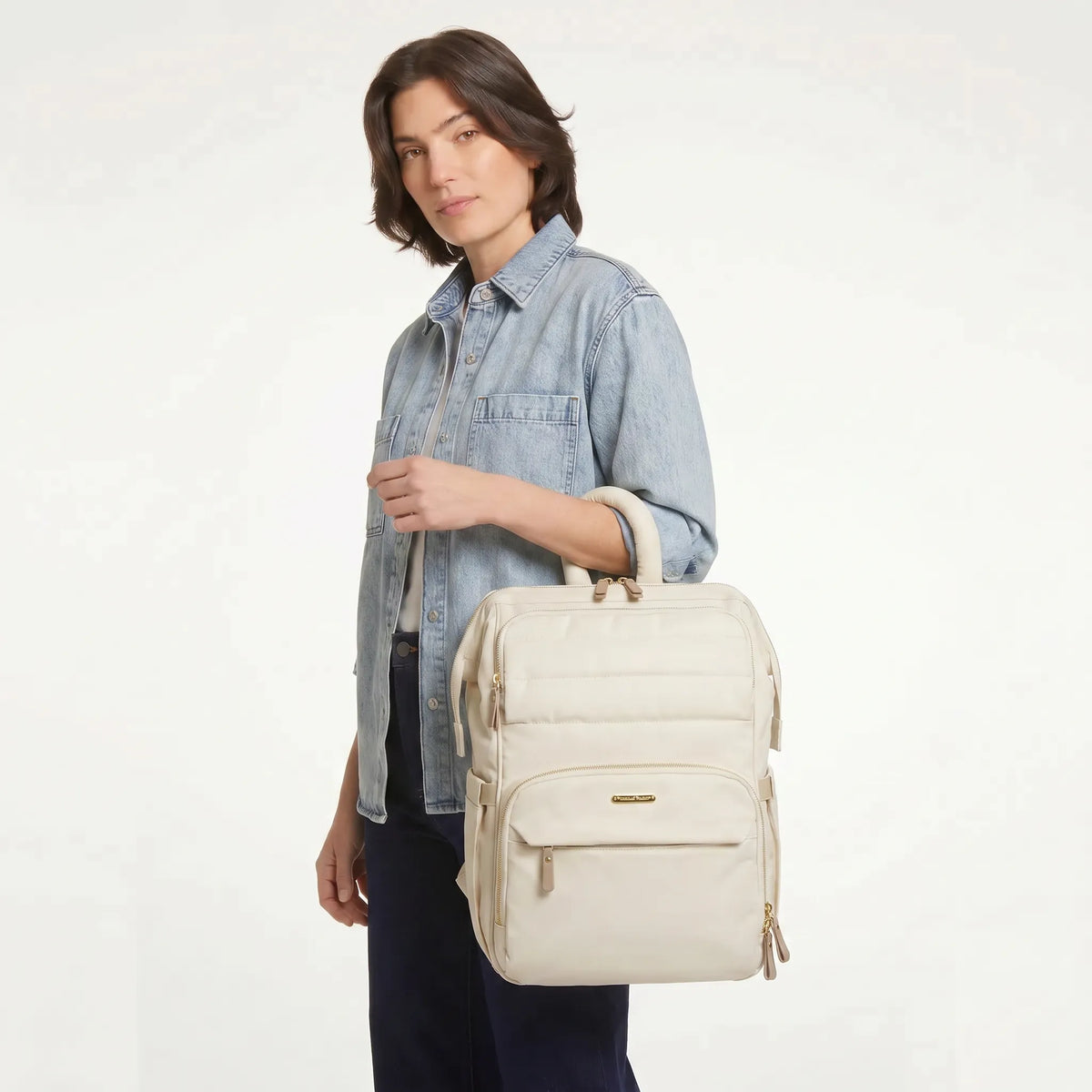 Jeune femme tenant à la main un sac à dos à langer crème monochrome avec hardware 
doré et poignée tubulaire rembourrée — photo lifestyle studio fond blanc
