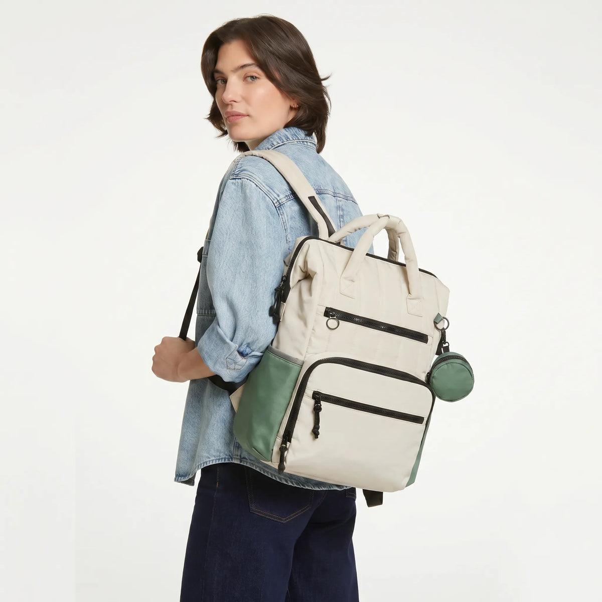 Femme aux cheveux courts bruns en chemise denim portant un sac à dos à langer crème avec détails vert sauge et pochette tétine ronde sur fond blanc studio