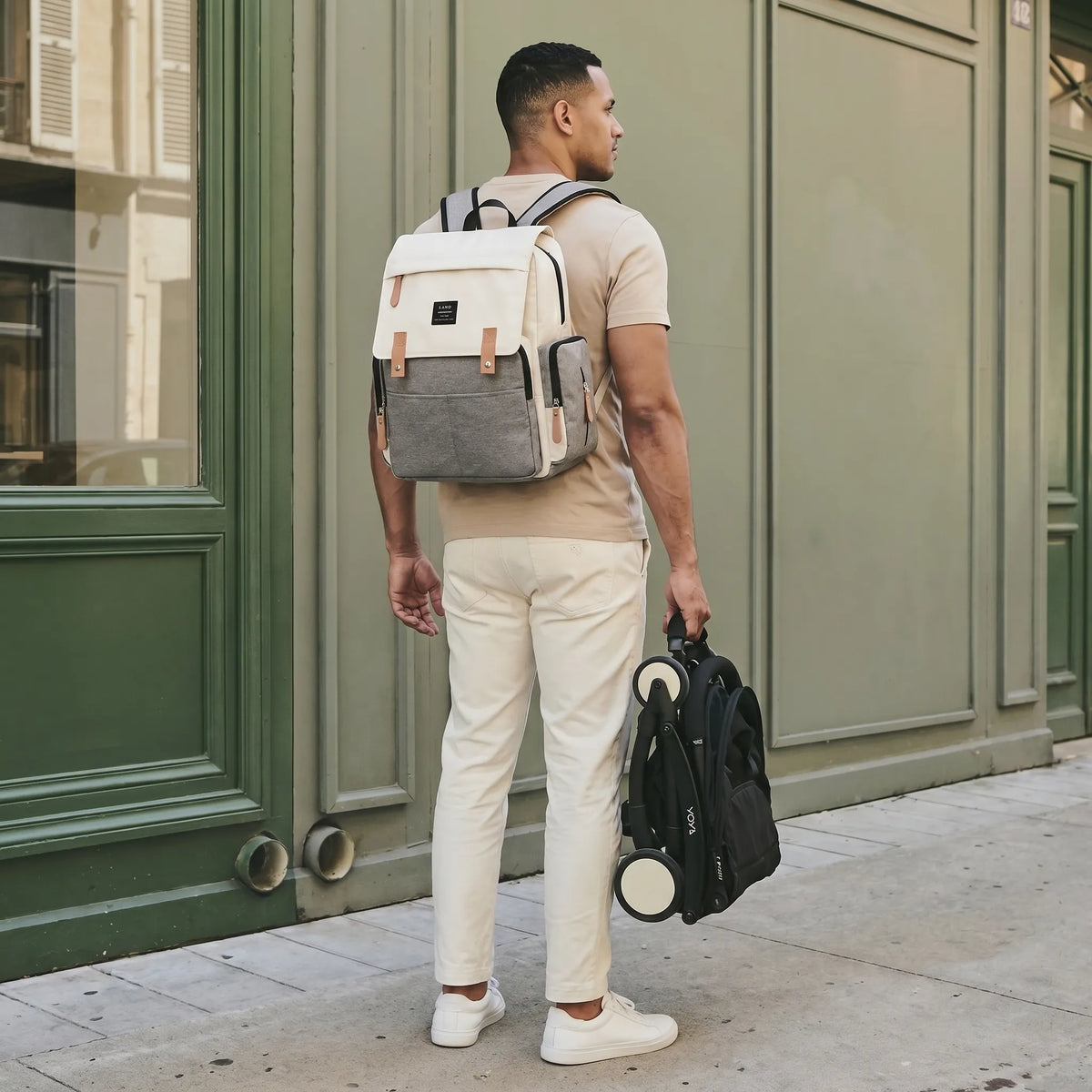 Papa de dos en t-shirt beige et pantalon crème portant un sac à dos à langer cream et gris color-block avec rabat à lanières cuir, tenant une poussette YOYO pliée à la main devant une devanture verte parisienne