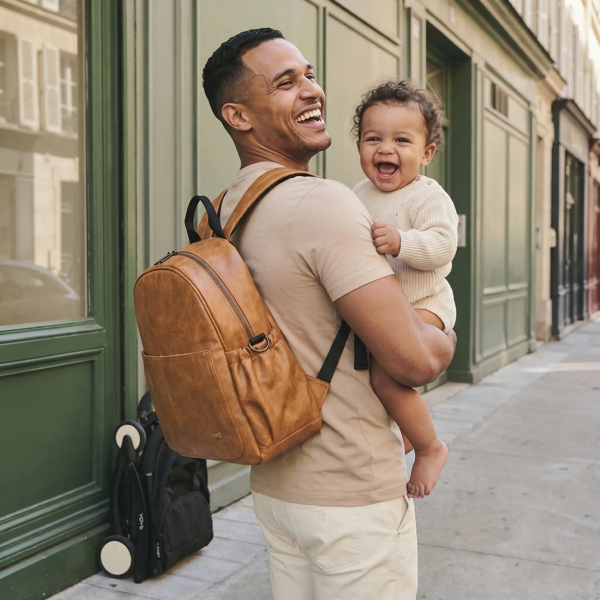 Papa souriant aux éclats portant son bébé métis aussi en plein éclat de rire sur la hanche, sac à dos à langer en cuir caramel vintage sur le dos, poussette YOYO pliée au sol, rue parisienne devanture verte