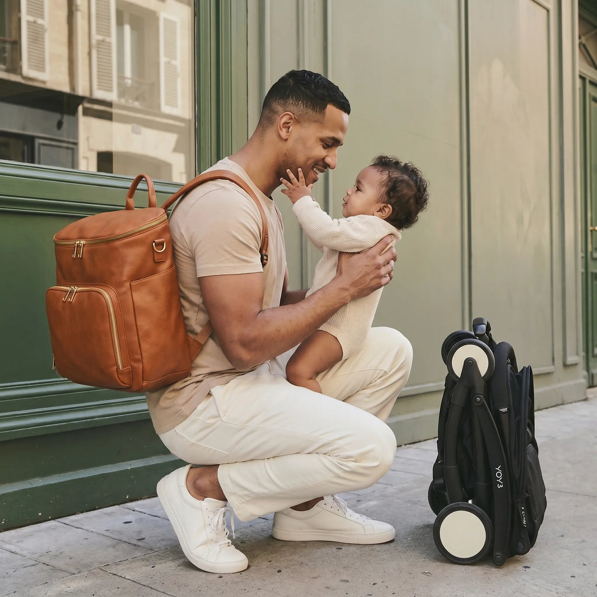 Papa souriant accroupi sur un trottoir parisien tenant son bébé métis dans ses bras avec un sac à dos à langer en cuir cognac sur le dos, dans un moment de complicité tendre, poussette YOYO pliée à côté