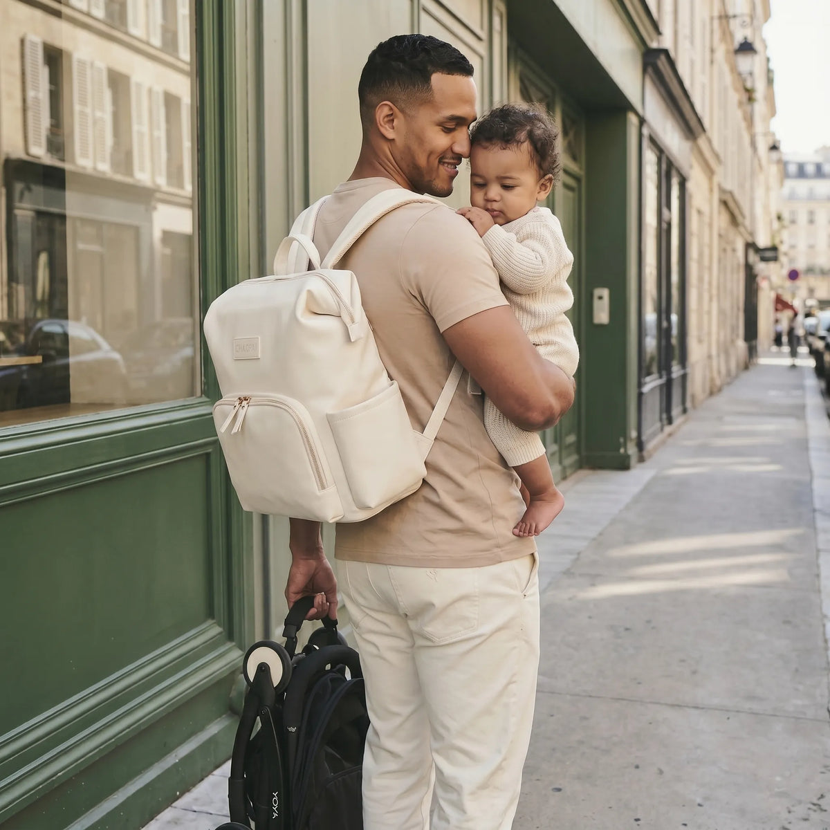 Papa souriant le front posé contre la tempe de son bébé métis dans une ruelle parisienne, portant un sac à dos à langer en cuir cream ivoire sur le dos et tirant une poussette YOYO derrière lui