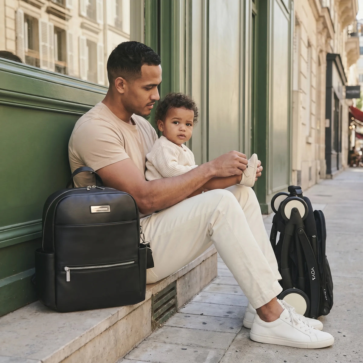 Papa assis sur un perron parisien en train de chausser tendrement son bébé métis assis sur ses genoux, sac à dos à langer en cuir noir posé à gauche et poussette YOYO pliée à droite
