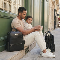 Papa assis sur un perron parisien en train de chausser tendrement son bébé métis assis sur ses genoux, sac à dos à langer en cuir noir posé à gauche et poussette YOYO pliée à droite