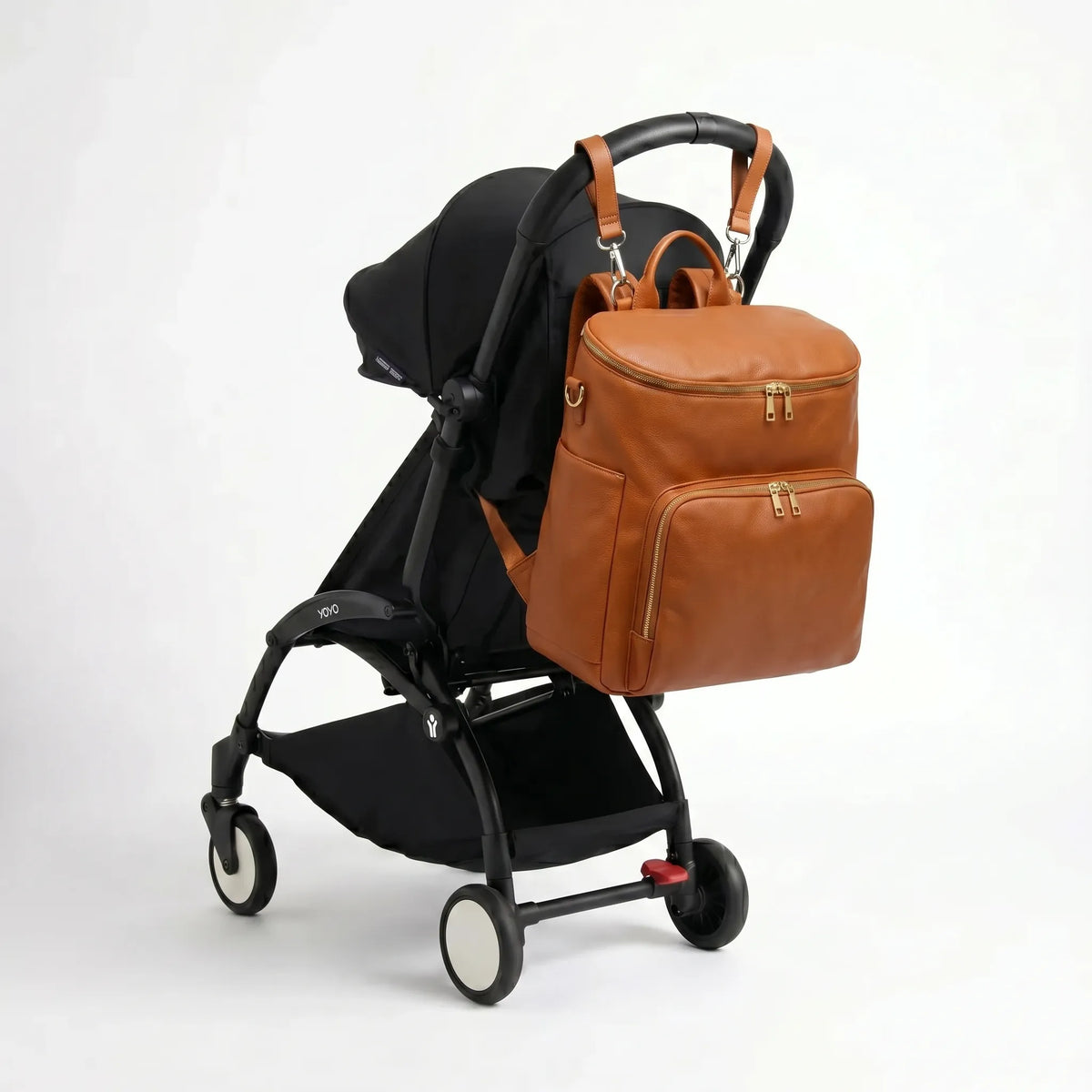 Sac à dos à langer en cuir PU camel avec zips dorés et sangles poussette accroché au guidon d'une poussette compacte noire de type Yoyo